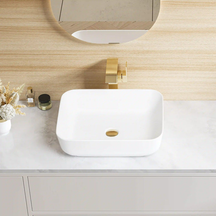 White rounded square ceramic vessel sink with brushed gold faucet and matching drain
